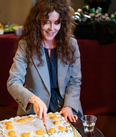 A woman with long curly hair wearing a gray blazer is smiling while cutting a decorated cake with orange slices on top. There's a drink in a glass beside her.