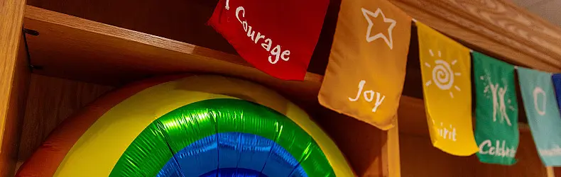 Colorful, rectangular flags with words like "Courage" and "Joy" hang on a wooden shelf. Below them, a partially visible, vibrant, round rainbow decoration is on the shelf.