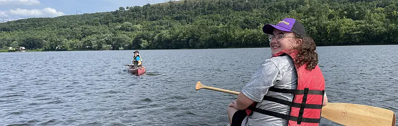 Two people canoeing on a calm lake surrounded by lush green hills. The person in the foreground wears a red life jacket and cap, smiling while holding a paddle. Another person is in the background paddling a red canoe.