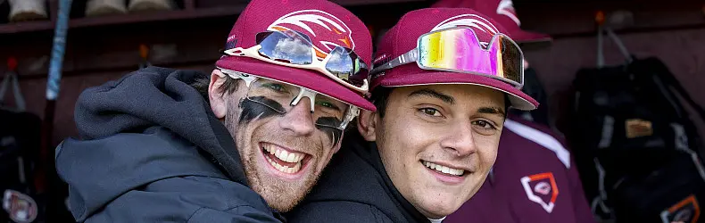 Two baseball players wearing maroon caps and uniforms smile at the camera. One has sunglasses and black eye paint. They appear to be in a dugout, with equipment visible in the background.