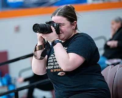 A person is taking a photo with a DSLR camera in a gymnasium. They are wearing a black t-shirt and standing near bleachers. Other people are visible in the background.