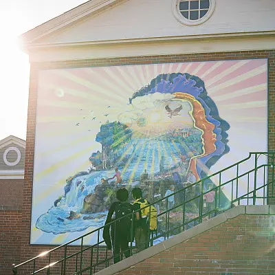 A large mural on a building wall shows a silhouette of a face filled with vibrant scenes of nature, including waterfalls, birds, and a bright sky. Three people stand at the bottom of a staircase, viewing the artwork. Sunlight shines from the top left.