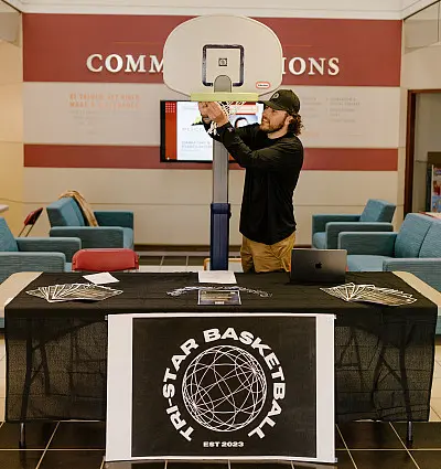 A person adjusts a mini basketball hoop on a table with a "Tri-Star Basketball" banner. The table is in a lobby area with seating and screens in the background.
