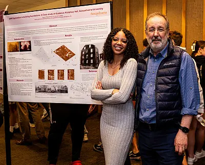 A woman in a gray dress and a man in a blue vest stand smiling in front of a research poster. The poster contains images and text, and is displayed at an indoor event with people in the background.