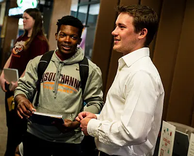 Two young men are talking and smiling indoors. One is wearing a gray hoodie and holding books, the other is in a white shirt. In the background, a person is standing with a laptop. The setting appears to be a casual or educational environment.