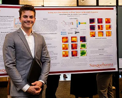 A person in a gray suit stands smiling in front of a research poster at Susquehanna University, featuring graphs and images related to "Testing Urns from Stability in Wind Tunnels". They hold a black folder.