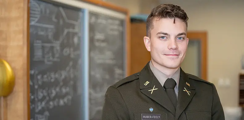 A man in military attire stands confidently in a room with chalkboards filled with equations in the background. He has short hair and a slight smile, and the setting appears to be academic or educational.