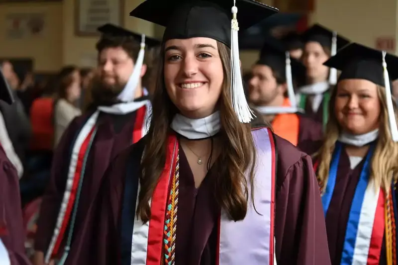 A group of graduates in maroon caps and gowns stand indoors, with a young woman in the foreground smiling. She is wearing honor cords and a white stole with red accents. Other graduates are visible in the background.