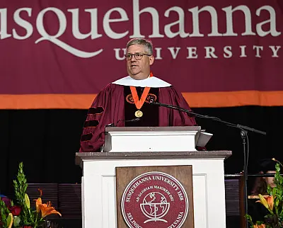 A speaker in academic regalia stands at a podium during a formal event at Susquehanna University. A large banner with the university's name hangs in the background.