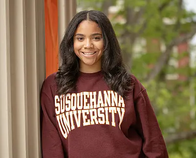 A person smiling while wearing a maroon sweatshirt with "Susquehanna University" written on it. They are standing outdoors, with green foliage visible in the background.