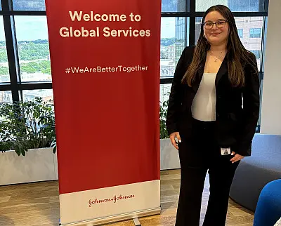A person in business attire stands beside a red banner with the text "Welcome to Global Services" and "#WeAreBetterTogether" in an office setting. Large windows and green plants are visible in the background.
