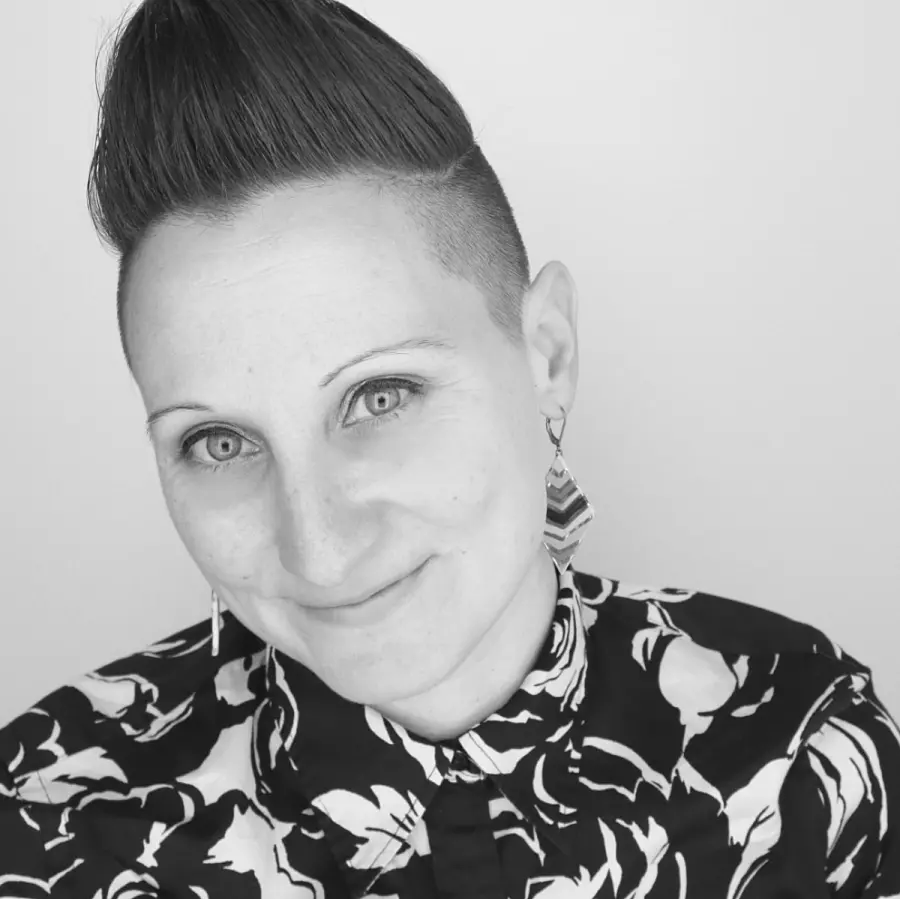 Person with short, styled hair and geometric earrings, wearing a patterned shirt, smiling softly against a plain background. Black and white image.