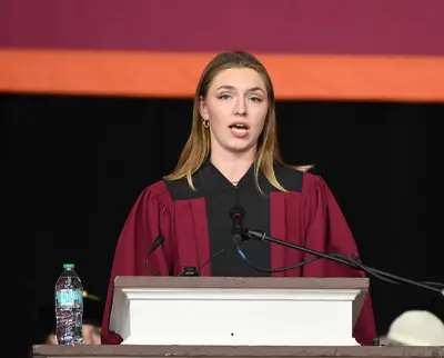 A person in a maroon graduation gown speaks at a podium with microphones. A water bottle sits on the podium, and a maroon and orange banner hangs in the background.