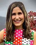 A woman with long brown hair smiles at the camera. She is wearing a colorful, polka-dotted sleeveless top. The background is slightly blurred, showing trees with red leaves under a cloudy sky.