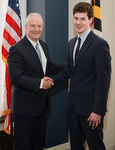 Two men in suits shaking hands and smiling in an office setting. An American flag is visible in the background. The man on the left has gray hair, and the man on the right has curly brown hair.