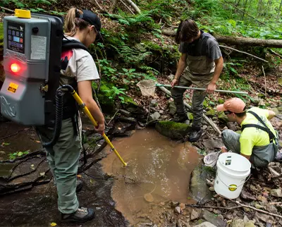 Three people in outdoor gear are standing by a small stream in a forest. One person holds a device on their back with a probe in the water, while the others hold nets and a bucket. They appear to be conducting water sampling or scientific research.