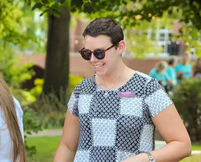 A person wearing sunglasses and a patterned black and white top smiles while standing outdoors. There is greenery in the background, and the person appears to be enjoying a sunny day.