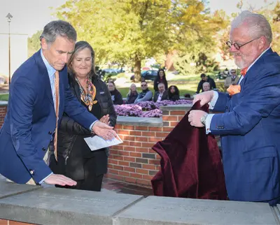 Three people stand near a brick wall outdoors; one man in a blue suit unveils a plaque, while another man and a woman observe. A small audience watches in the background amidst autumn trees.