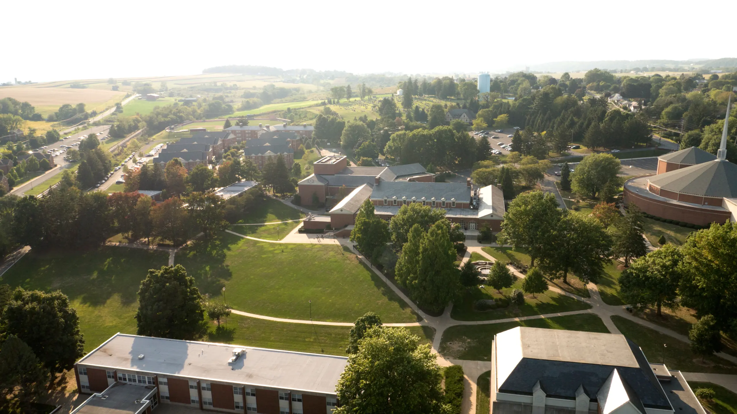 Aerial view of a sprawling campus with multiple buildings surrounded by lush green trees and open grassy areas. Pathways connect the buildings, creating a serene and structured layout. A water tower is visible in the background.