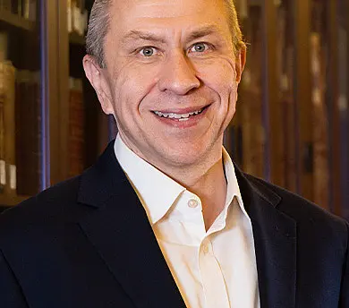 A smiling man in a black suit and white shirt stands in a library with wooden bookshelves filled with books in the background.