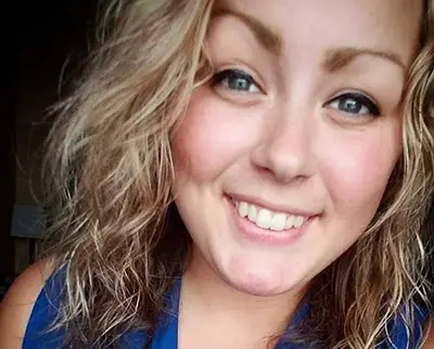 A person with wavy, light brown hair smiles at the camera. They are wearing a blue shirt. The background is softly lit.