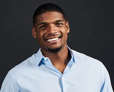 Smiling person with short hair, wearing a light blue button-up shirt, against a dark background.