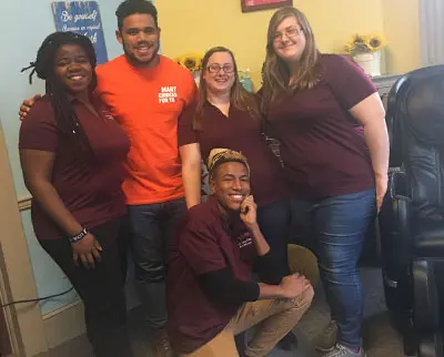 A group of five people smiling and posing together indoors. Four are wearing maroon shirts, and one is in an orange shirt. They stand and kneel in front of a wall decorated with sunflowers and motivational signs.