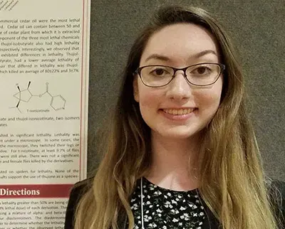 A person with long hair and glasses smiling in front of a scientific poster featuring chemical diagrams and text.