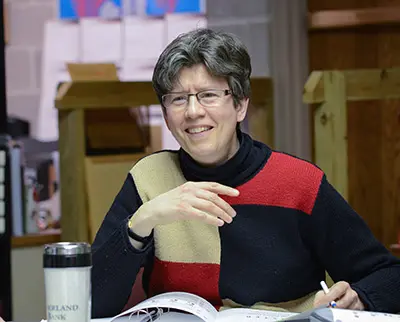 A person with short gray hair is sitting at a table, smiling and holding a pen. They are wearing glasses and a sweater with red, black, and beige patches. A travel mug and an open book are on the table.