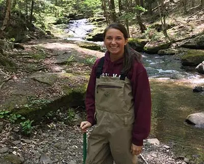 A person wearing waders and a maroon hoodie stands by a rocky stream in a forest. The background features flowing water, rocks, and greenery under a canopy of trees.