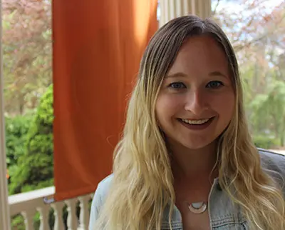 A person with long blonde hair smiles while standing outside in front of a porch with white columns. There is an orange banner behind them and greenery in the background. They are wearing a light-colored jacket.