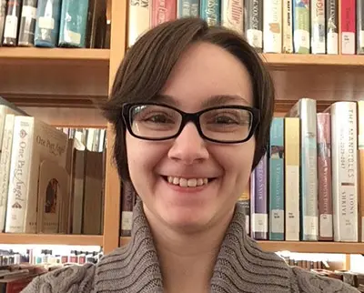 A person with short brown hair and glasses is smiling in front of a bookshelf filled with various books. They are wearing a gray sweater.