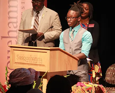 A young individual speaks at a podium labeled "Susquehanna University" during a formal event. Two people stand behind, one holding papers. The setting appears academic or ceremonial, with decorative elements in the foreground.