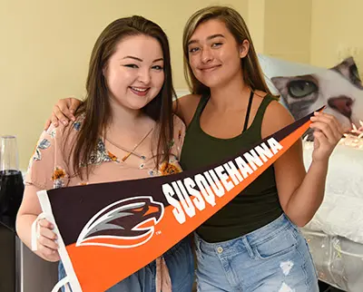 Two smiling young women stand close together, holding a Susquehanna University pennant. One wears a floral top, the other a green tank top and denim shorts. A pillow with a cat design is visible in the background.