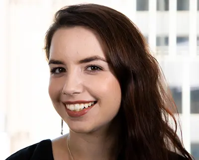 A woman with long dark hair smiles at the camera against a blurred office window background. She is wearing a black top and earrings.