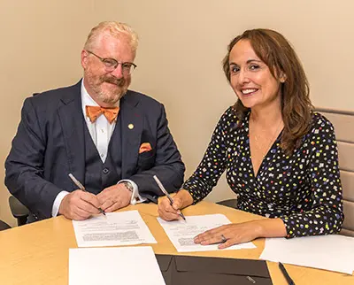 A man in a suit with an orange bow tie and a woman in a polka dot dress smile while signing documents at a table.