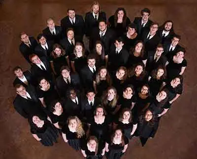 A group of people dressed in black formal attire stand closely together, forming a heart shape. They are looking up at the camera, which captures them from above. The background is a simple brown surface.
