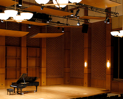 A concert hall with a grand piano on a wooden stage. The room features wood paneling and wall sconces, with a large suspended acoustic panel overhead. Rows of seating are partially visible in the foreground.