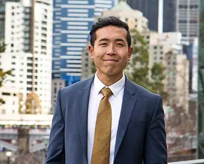 A person in a suit and tie stands outdoors with a city skyline in the background. They have short dark hair and are smiling slightly, with tall buildings visible behind them on a clear day.