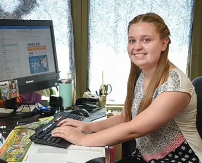 A woman seated at a desk, typing on a keyboard with a cheerful expression. She is in an office setting with a computer screen displaying a website, various office supplies, and sunlight streaming through the window behind her.