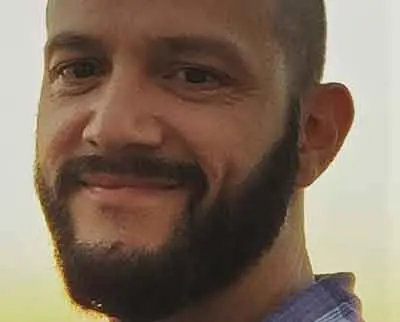 A man with a beard smiles at the camera. He is wearing a blue and white checkered shirt. The background is softly blurred, suggesting an outdoor setting.