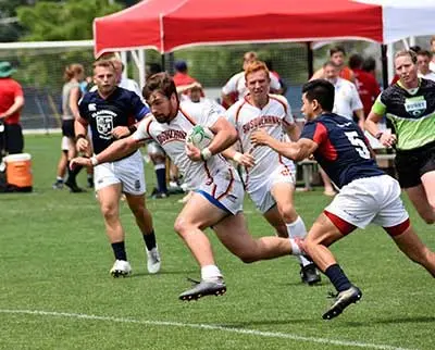 A rugby player in white, mid-stride holding the ball, is pursued by opponents in dark jerseys. The game is set on a grassy field with a red tent in the background. Other players and spectators are visible.