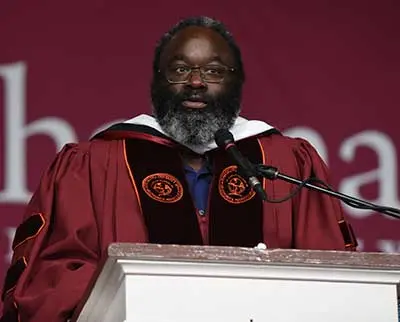 A person wearing academic regalia and glasses stands at a podium with a microphone. The background features a maroon backdrop with partial white lettering visible.