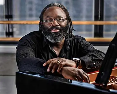 A man with glasses and a beard is seated at a piano, smiling slightly. He is wearing a dark shirt, and the background shows blurred windows and a railing.