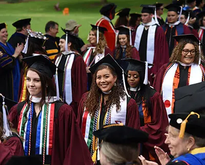Graduates in maroon caps and gowns walk in a procession outdoors. They wear colorful stoles and cords. Faculty members in academic regalia stand on either side, clapping and smiling. The setting is festive and celebratory.