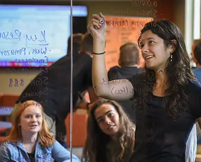 A woman writes on a transparent board while three other people sit around her, engaged. Various notes and diagrams are visible on the board. The background shows an office setting with screens displaying information.