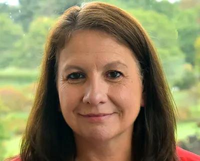 A woman with long brown hair is smiling gently at the camera. She is wearing a red top. The background shows a blurred, lush green scenery, suggesting an outdoor setting on a bright day.