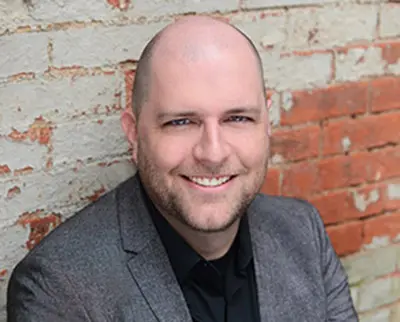 A smiling person with a shaved head is wearing a gray blazer and black shirt. They are seated in front of a brick wall with peeling paint.