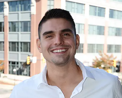 A man with short dark hair and a goatee is smiling while wearing a white collared shirt outside. A blurred urban background with a modern building and trees is visible behind him.