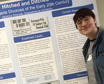 A person stands next to a research poster titled "Hitched and Ditched: Pennsylvania Divorces of the Early 20th Century." The poster includes sections on examined cases and current research. The person is smiling and wearing glasses and a striped shirt.